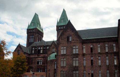 Historic brick building with green-topped towers under a cloudy sky, surrounded by trees.
