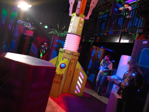 A colorful laser tag arena with players in futuristic gear, featuring a large, vibrant structure in the center.