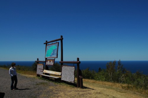A person stands near an informational sign overlooking a vast blue ocean under a clear sky.