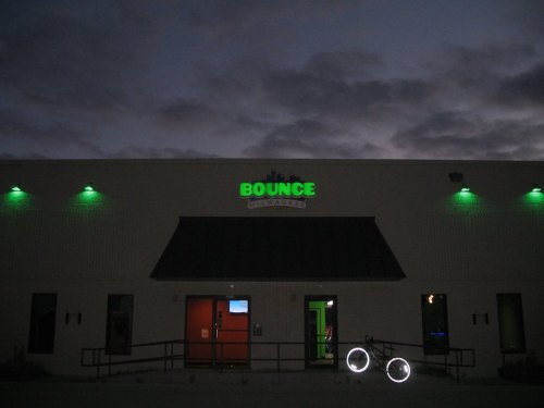 A dimly lit building with a green "BOUNCE" sign, featuring a ramp and two bicycles with illuminated wheels.