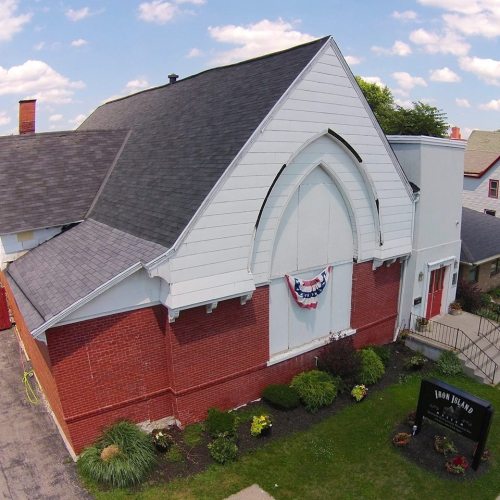 Aerial view of a building with a peaked roof, red brick walls, and decorative bunting on the entrance. Green landscaping surrounds it.
