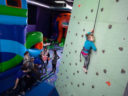 A child climbs a rock wall in a colorful indoor play area with inflatable structures and a group of people nearby.