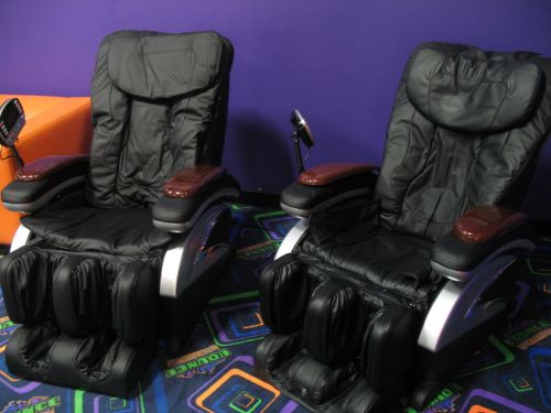 Two black massage chairs with wooden armrests, set against a purple wall and colorful patterned carpet.