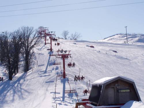 Ski lift chairs ascend a snowy hill under a clear blue sky, with trees lining the slope.