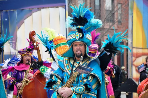 A performer in vibrant blue and feathered costume holds a saxophone, surrounded by others in colorful attire at a parade.
