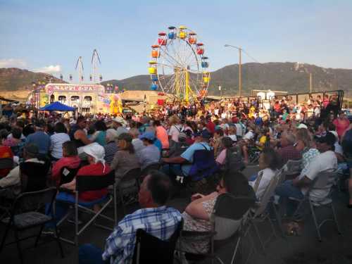 A large crowd sits in folding chairs at a fair, with a Ferris wheel and carnival rides in the background.