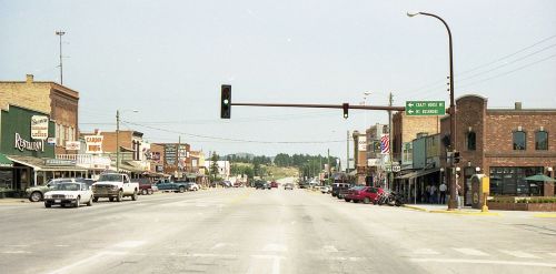 A small town street with shops, restaurants, and traffic lights, featuring parked cars and pedestrians along the sidewalks.
