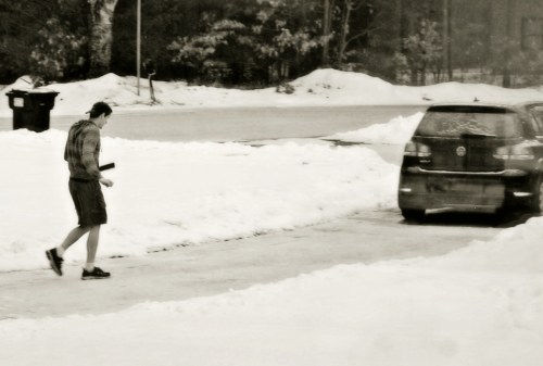 A person in shorts walks on a snowy driveway beside parked cars.