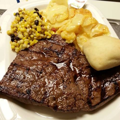 A plate featuring a grilled steak, corn and black bean salad, cheesy potato slices, and a dinner roll.