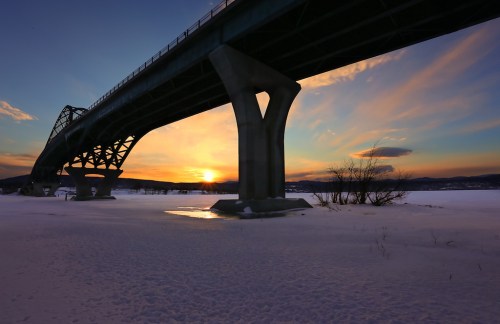 A bridge spans over a snowy landscape at sunset, with vibrant colors in the sky and shadows cast by the structure.