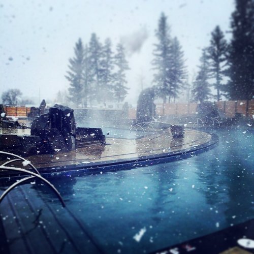 A snowy scene featuring a misty pool surrounded by wooden decking and tall evergreen trees.