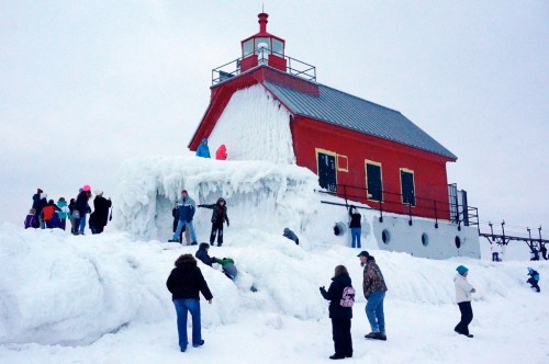 A red lighthouse covered in ice, with people playing in the snow around it on a cold winter day.