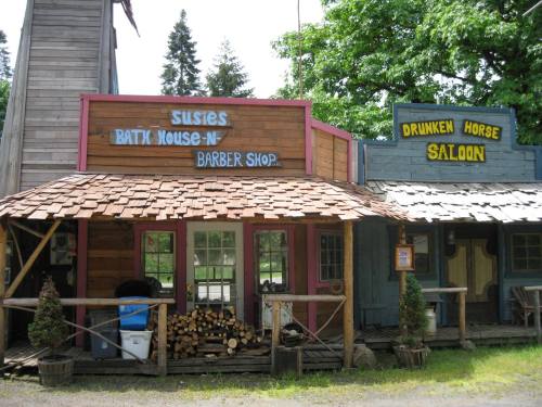 A rustic storefront featuring "Susie's Batik House-N Barber Shop" and "Drunken Horse Saloon" signs.