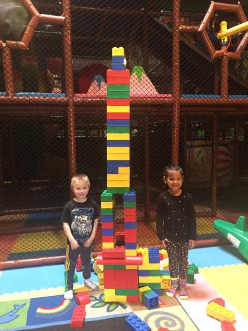 Two children stand beside a tall tower made of colorful building blocks in a play area.