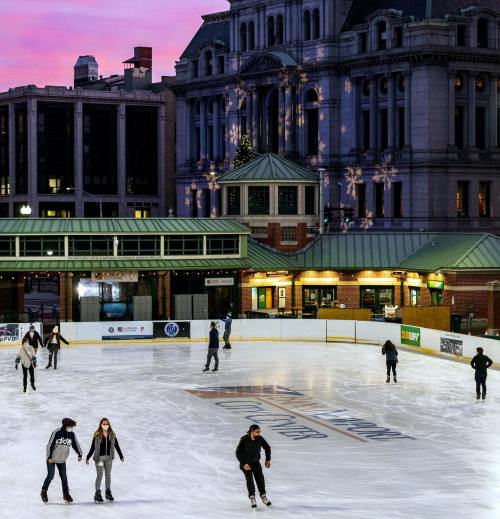 People ice skating on a rink with a cityscape and colorful sunset in the background.