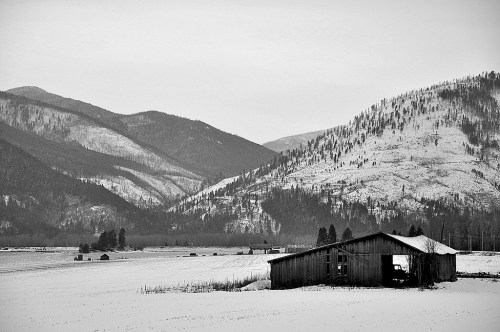A weathered barn sits in a snowy landscape, surrounded by mountains and trees, in a serene black-and-white scene.