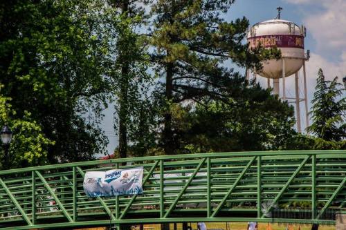 A green pedestrian bridge with a banner, surrounded by trees, and a water tower in the background.