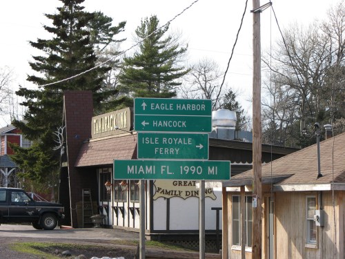 Signpost indicating directions to Eagle Harbor, Hancock, Isle Royale Ferry, and Miami, FL, with a restaurant in the background.