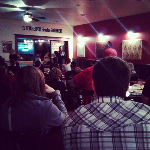 A comedian performs on stage in a dimly lit venue, with an audience seated at tables watching attentively.