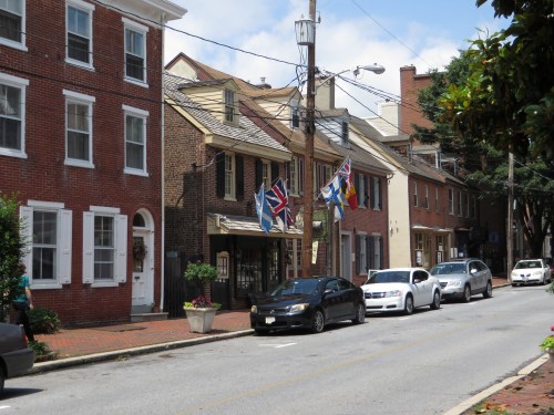 A charming street lined with historic brick buildings and flags, featuring parked cars and a clear blue sky.