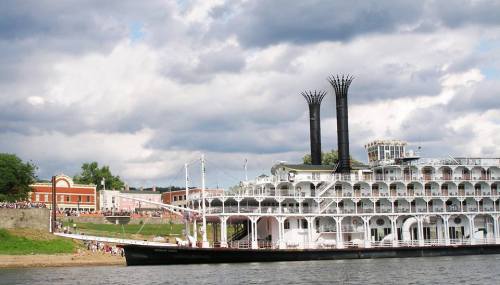A large steamboat with two tall smokestacks docked by a riverside, under a cloudy sky.