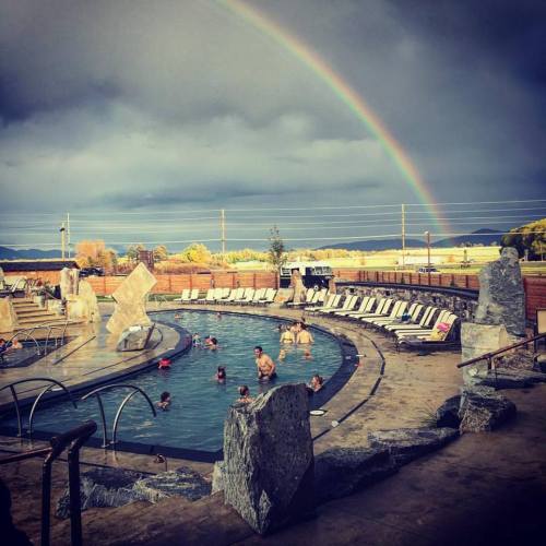 A scenic outdoor pool with people swimming, surrounded by rocks, under a rainbow and cloudy sky.