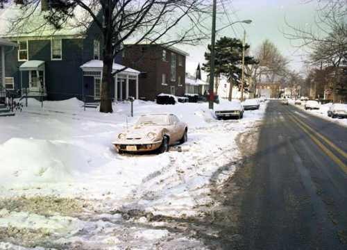 A snowy street scene with parked cars and a vintage car partially buried in snow beside residential buildings.