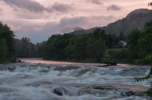 A serene river flows through a lush landscape at dusk, with mountains and a house in the background under a cloudy sky.