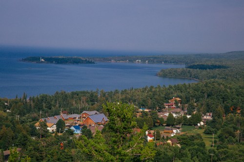 A scenic view of a coastal town surrounded by lush greenery and a calm blue lake under a hazy sky.