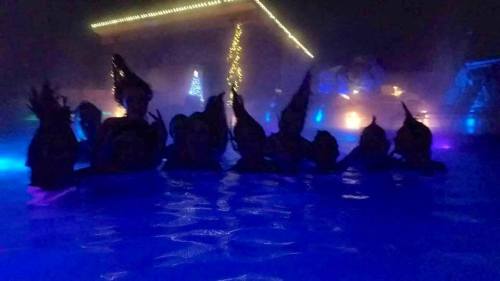 A group of people silhouetted in a brightly lit pool at night, surrounded by colorful lights and mist.
