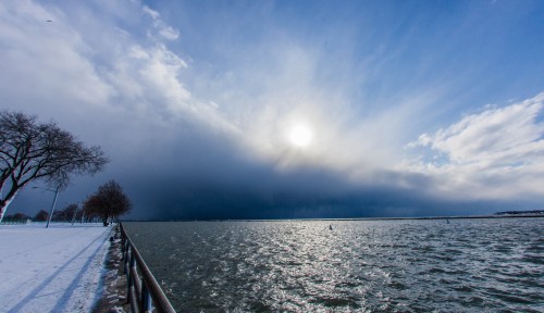 A serene lakeside view with dark clouds, sunlight peeking through, and snow-covered trees along the shore.