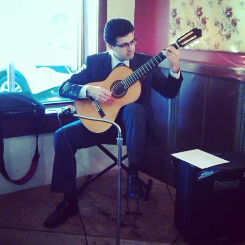 A musician in a suit plays a guitar while seated in a cozy indoor setting.