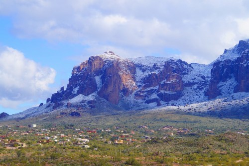 Snow-capped mountains rise above a green valley dotted with small houses under a partly cloudy sky.