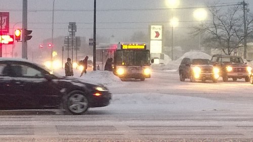 A snowy intersection with a bus and cars, as pedestrians navigate through the winter weather.
