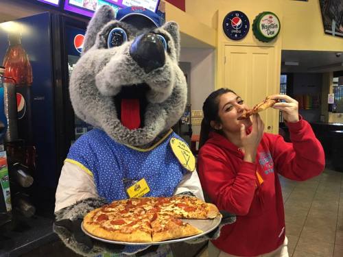 A person in a red hoodie enjoys a slice of pizza next to a mascot holding a pizza tray in a restaurant.