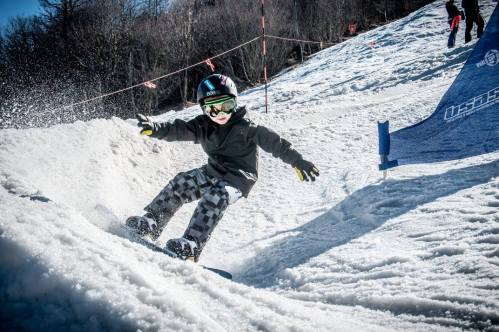 A young child snowboarding on a snowy slope, wearing a helmet and goggles, with snow spraying around them.