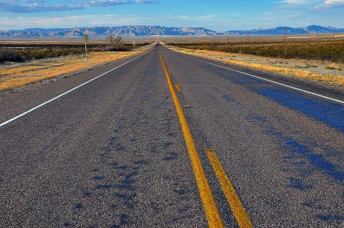 A long, empty road stretches into the distance, flanked by grasslands and mountains under a clear blue sky.