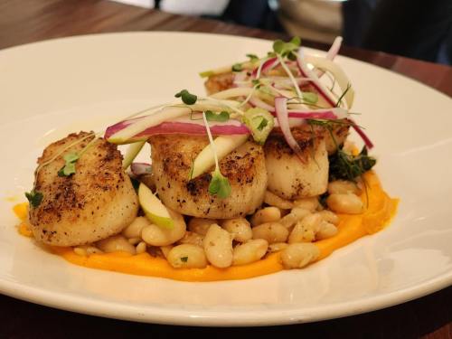 Seared scallops on a bed of white beans and sweet potato puree, garnished with microgreens and sliced vegetables.