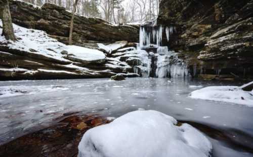 A serene winter scene featuring a frozen waterfall surrounded by snow-covered rocks and a still icy pond.