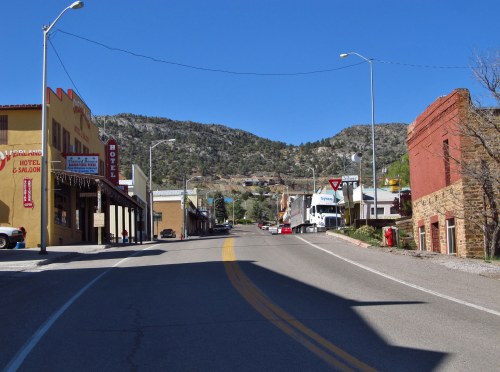 A quiet street in a small town, lined with buildings and mountains in the background under a clear blue sky.