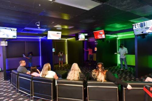 A group of people in a dimly lit room with colorful lights, engaging with virtual reality headsets and screens.