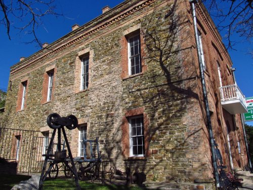Historic stone building with a shadow of a tree, featuring a balcony and outdoor seating area. Clear blue sky above.
