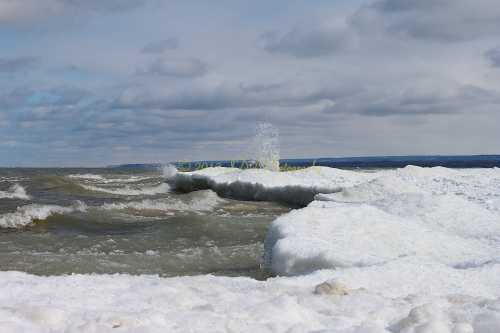 Waves crash against icy shores under a cloudy sky, creating splashes of water amidst the frozen landscape.