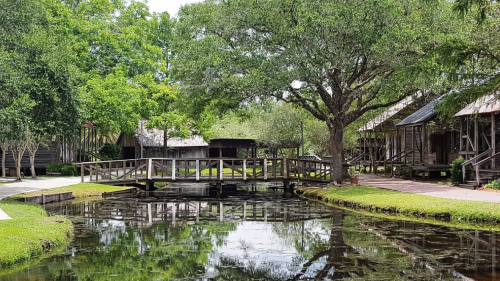 A serene scene featuring a wooden bridge over a calm pond, surrounded by lush greenery and rustic buildings.