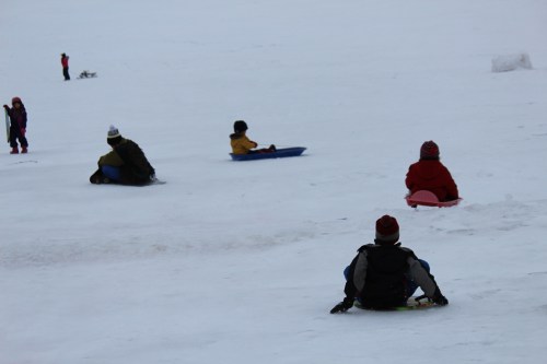 Children sledding down a snowy hill, with a few others playing in the background.