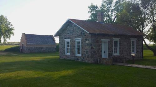 Two historic stone buildings on a grassy landscape, surrounded by trees and open fields under a clear sky.