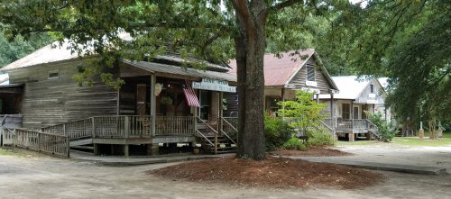 A row of rustic wooden buildings with porches, surrounded by trees and a dirt path.