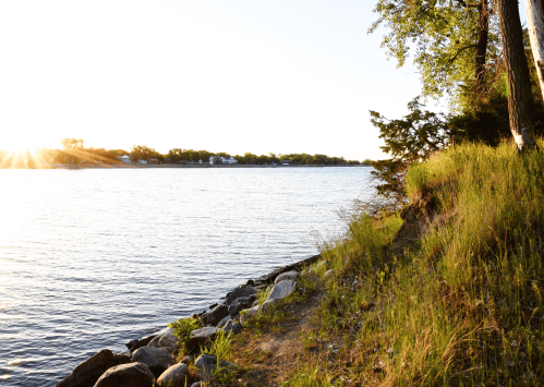A serene riverside scene at sunset, with grassy banks and rocks along the water's edge.