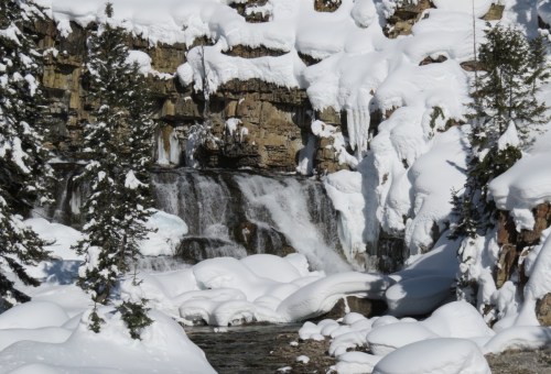 A snowy landscape featuring a waterfall cascading over rocks, surrounded by snow-covered trees and icy formations.