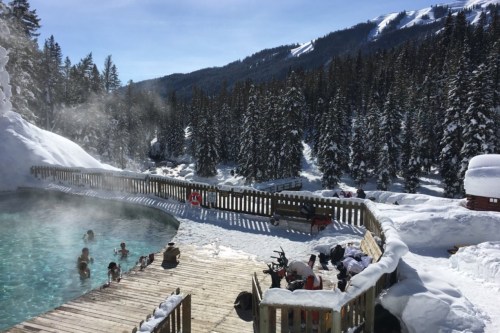 A scenic hot spring surrounded by snow-covered trees and mountains, with people enjoying the warm water.